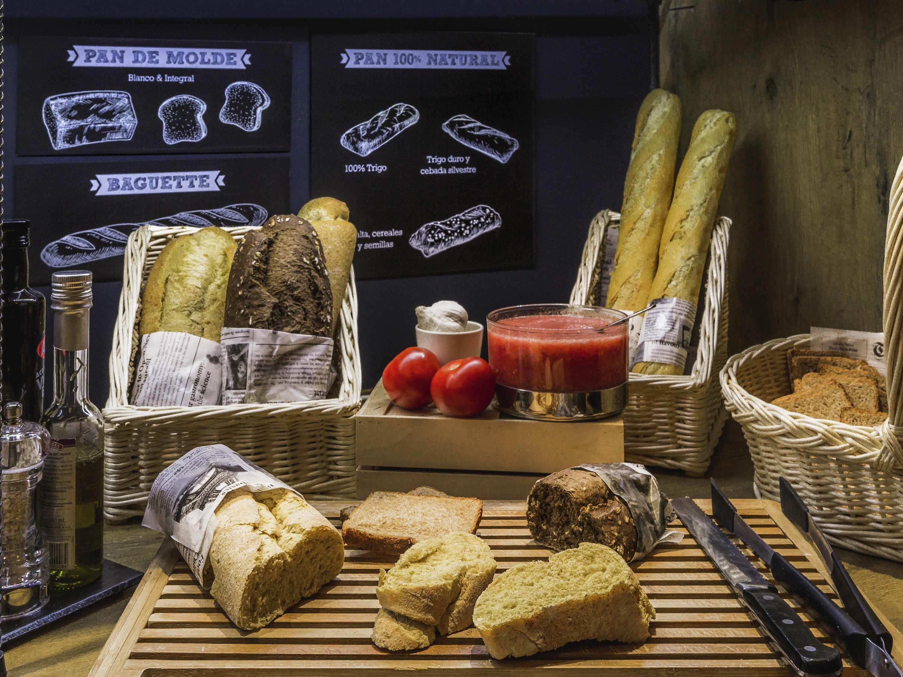 A variety of breads and pastries displayed on a wooden table with baskets, a glass of tomato sauce, and a knife.