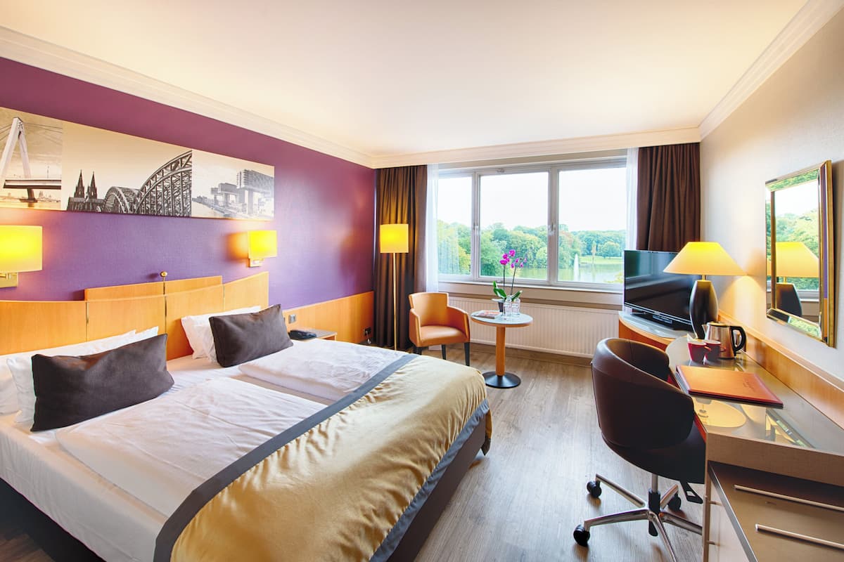 Quarto de hotel com uma cama grande, uma escrivaninha com computador, uma cadeira e janela com vista para vegetação.