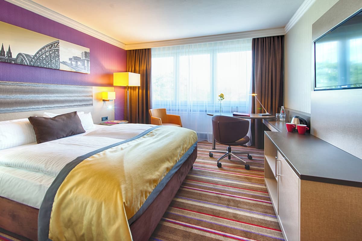 Quarto de hotel com uma cama grande, uma escrivaninha, uma cadeira, uma televisão e uma janela com cortinas.