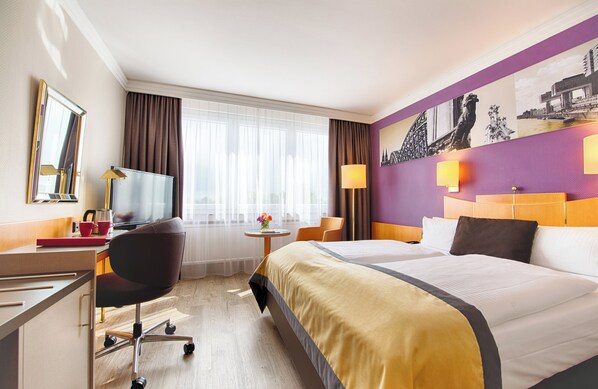 Una habitación de hotel con una cama grande, un escritorio con una computadora, una silla, una mesita con un florero y una ventana con cortinas.