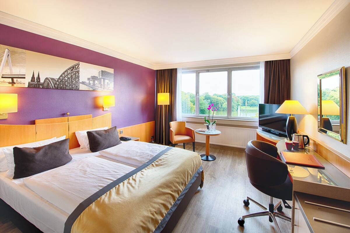 Ein Hotelzimmer mit einem großen Bett, einem Schreibtisch mit Computer, einem Stuhl und einem Fenster mit Blick auf Grünfläche.