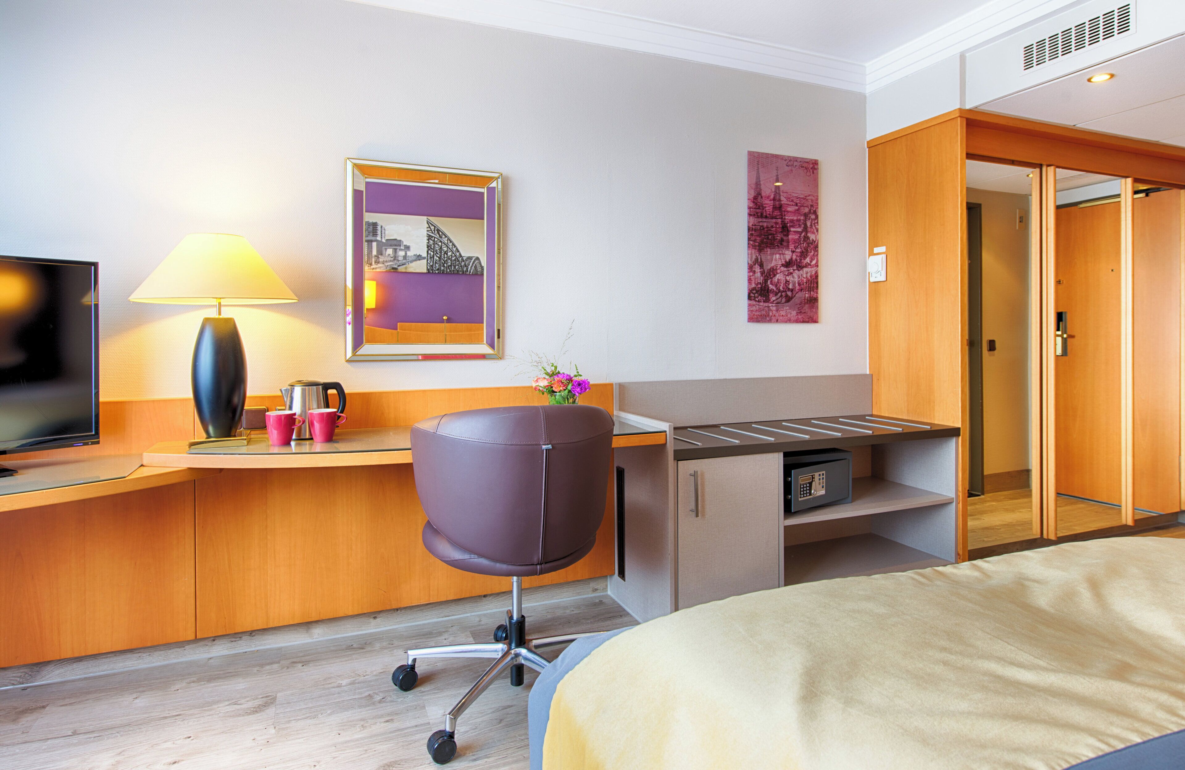 Habitación de hotel con cama, escritorio con lámpara, una silla, televisor y obras de arte en las paredes.