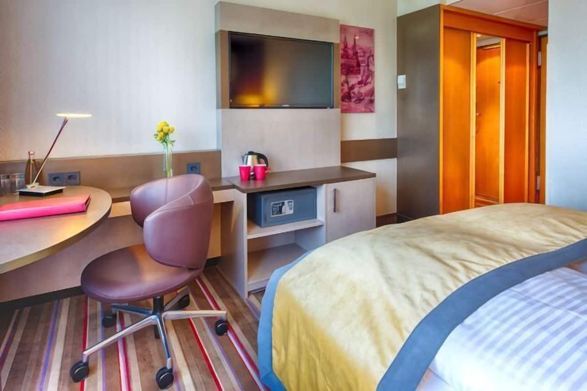 Quarto de hotel com cama, escrivaninha, cadeira, televisão e guarda-roupa.