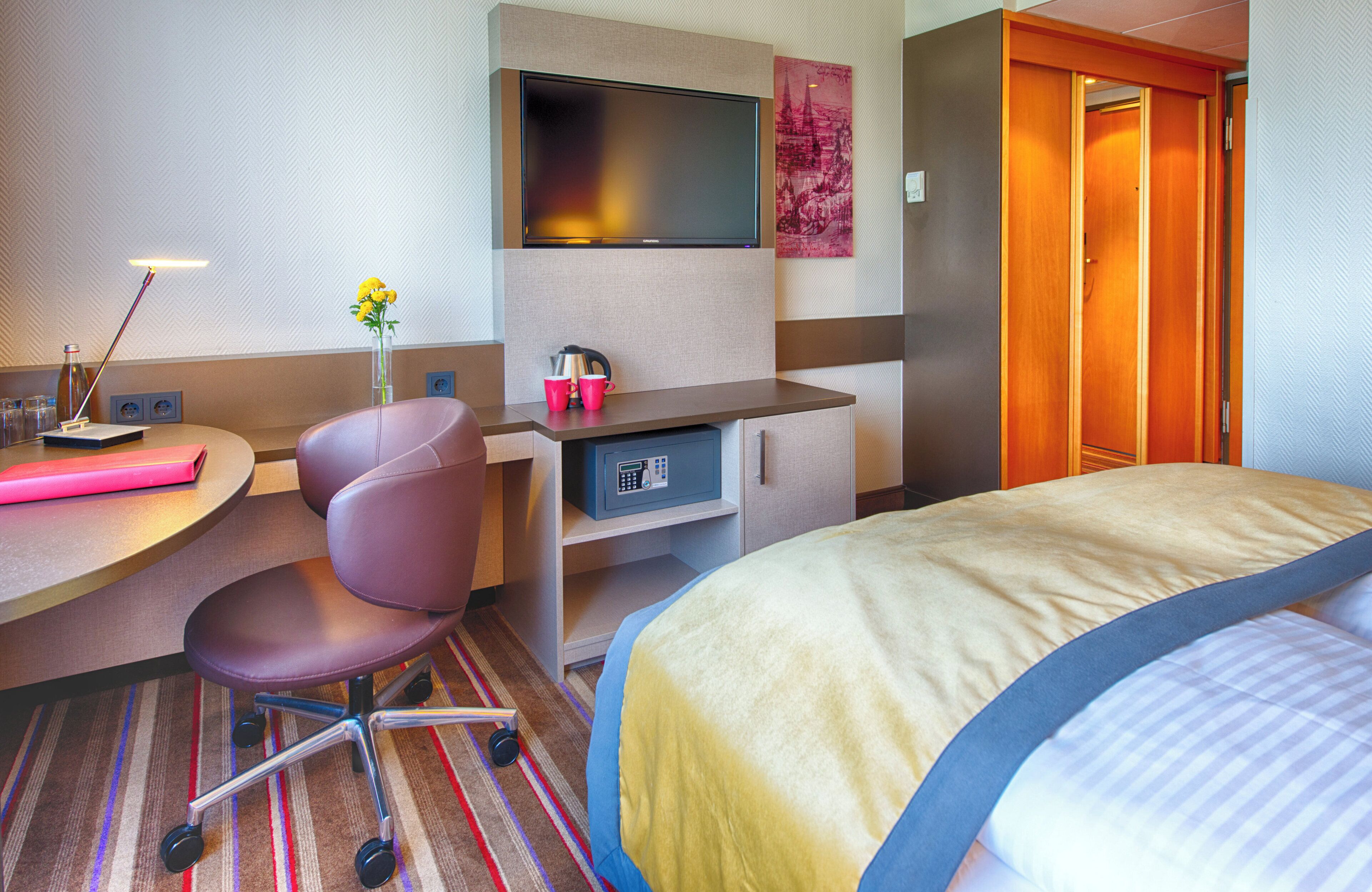 A hotel room with a bed, desk, chair, TV, and a safe.
