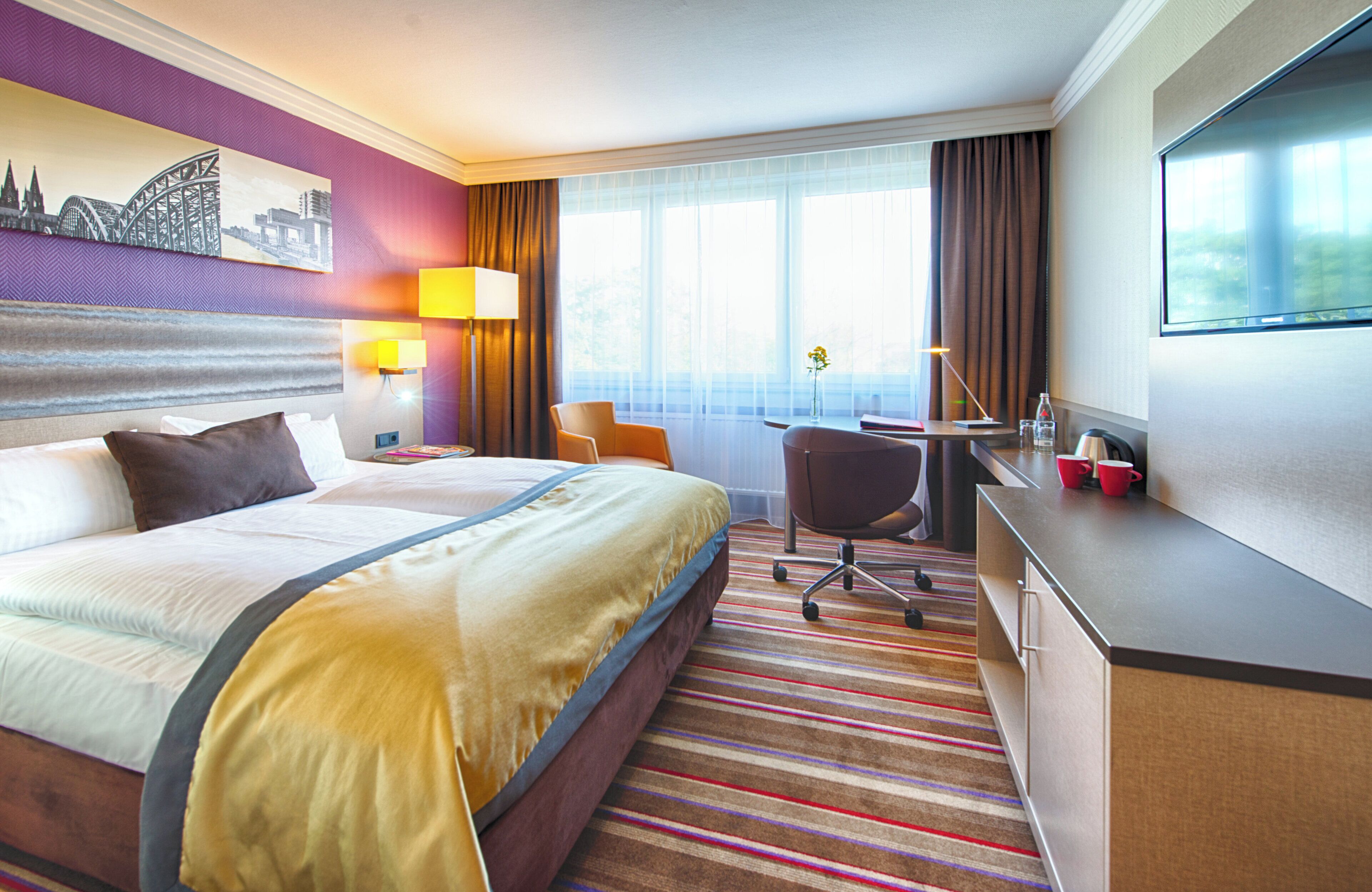 A hotel room with a large bed, a desk, a chair, a TV, and a window with curtains.
