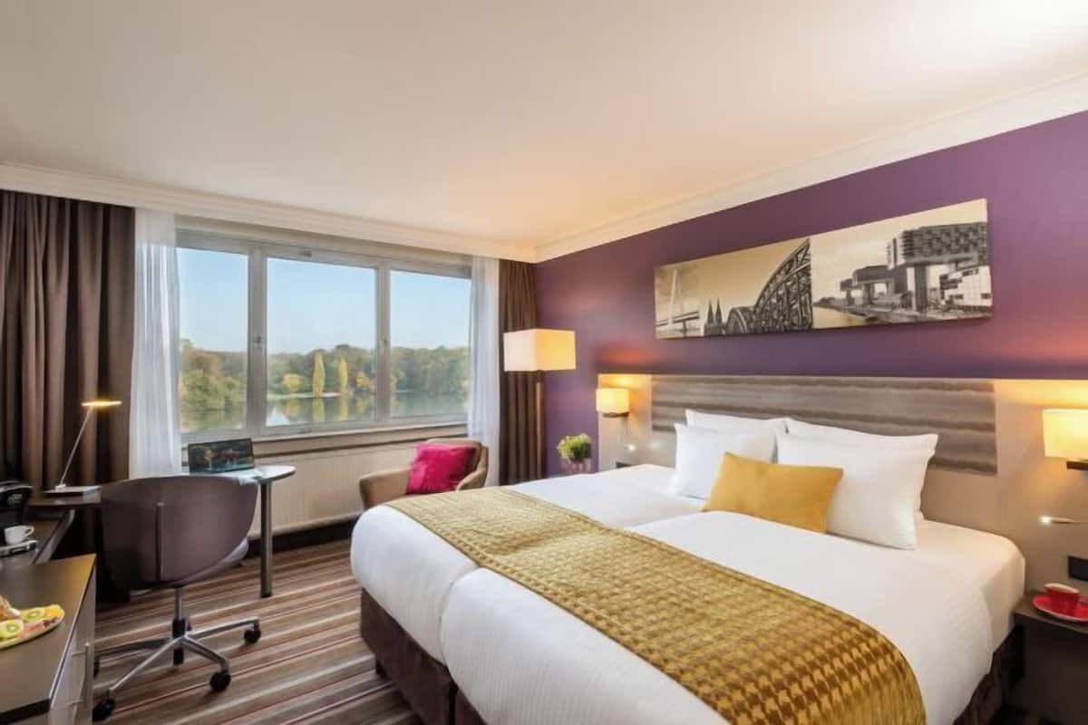 Quarto de hotel com uma cama grande, uma escrivaninha, uma cadeira, uma janela com vista e um quadro na parede.