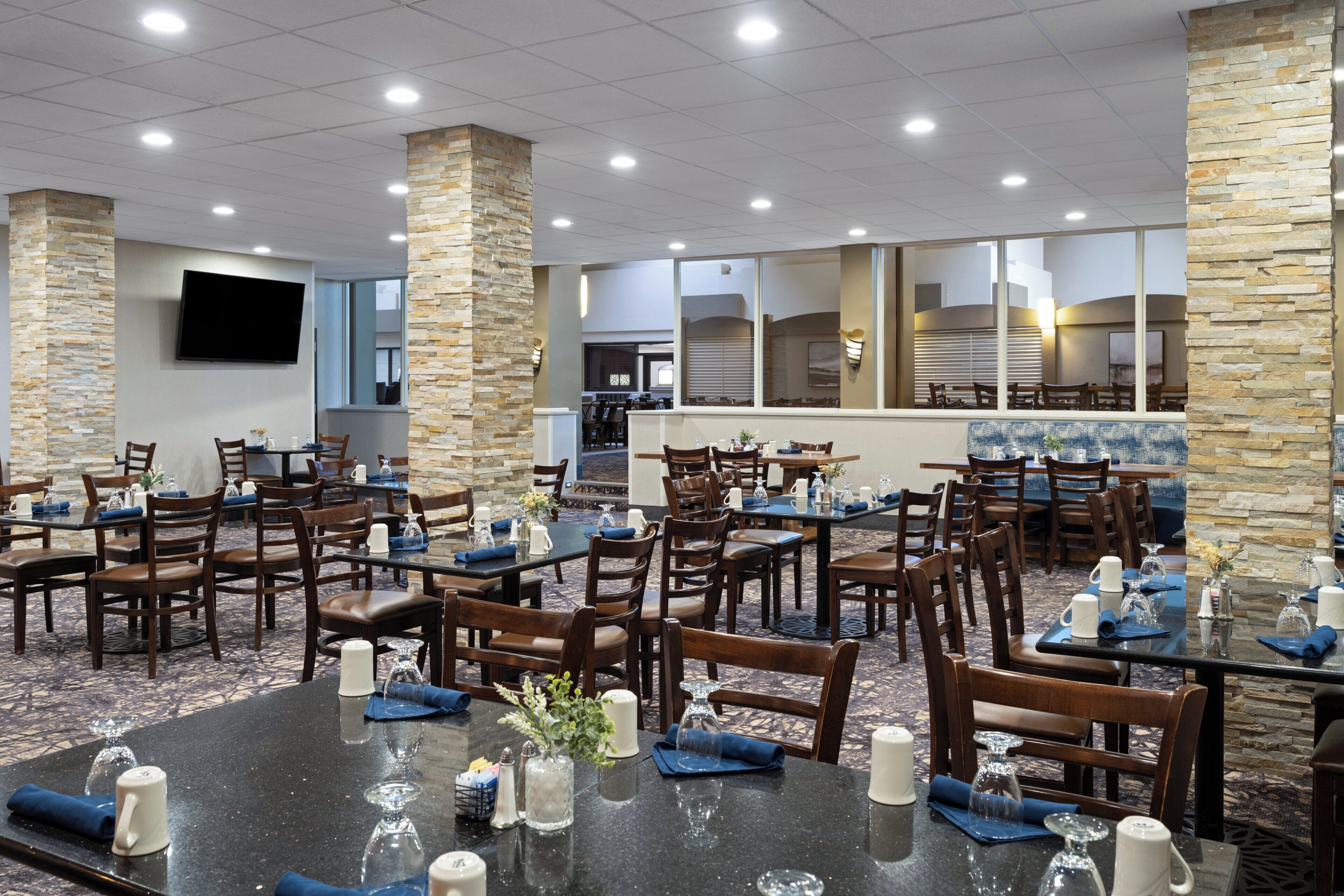 Un espace de restauration d’hôtel comprenant plusieurs tables et chaises, des piliers en pierre et un téléviseur à écran plat fixé au mur.