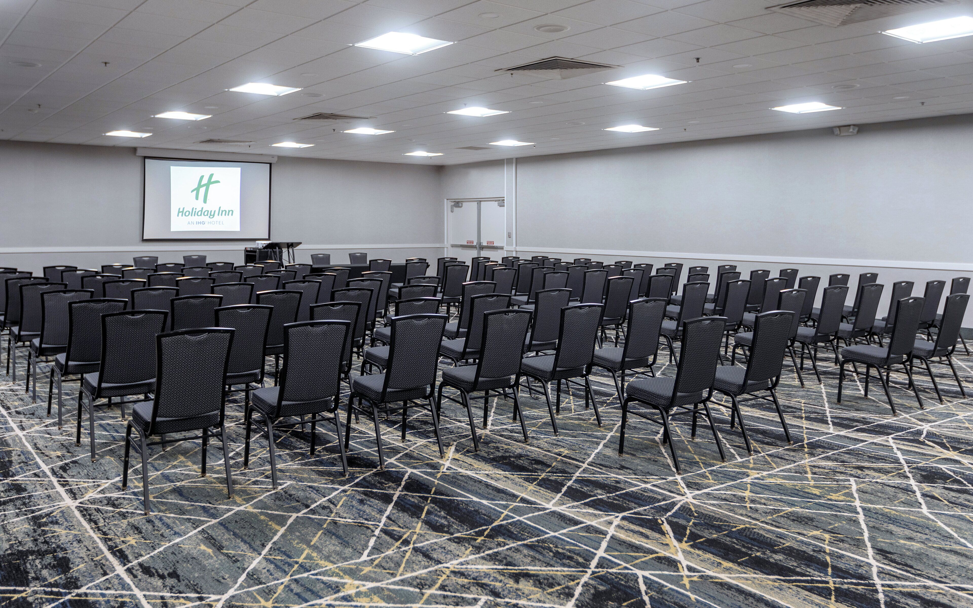 A conference room with rows of chairs, a projector screen, and a carpeted floor.