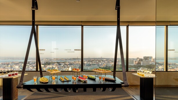 Habitación en un rascacielos con vistas panorámicas de la ciudad, una mesa de buffet con comida y bebidas variadas, y un amplio ventanal con vistas al paisaje urbano.
