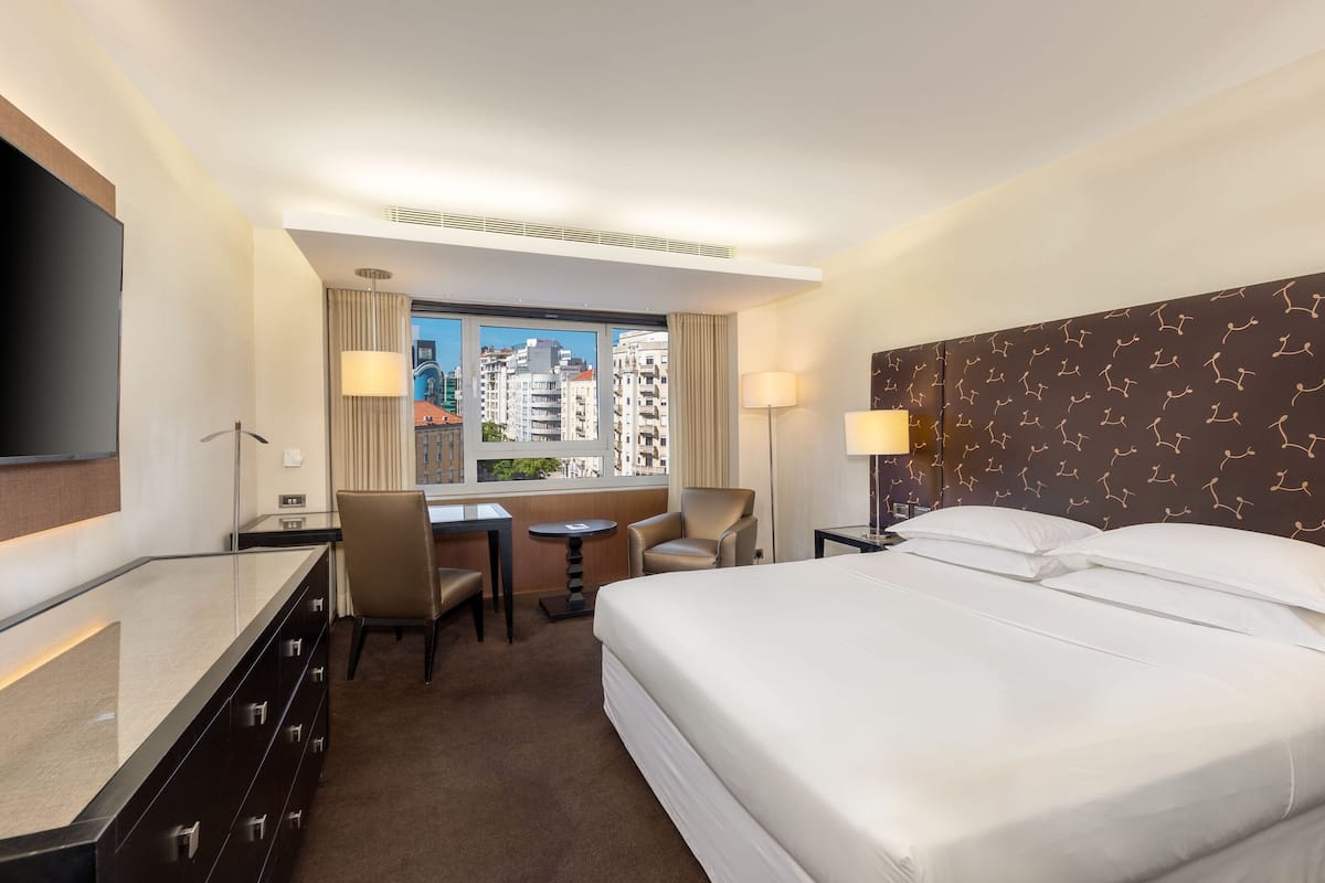 Habitación de hotel con una cama grande, un escritorio con silla, televisión y ventana con vista a la ciudad.