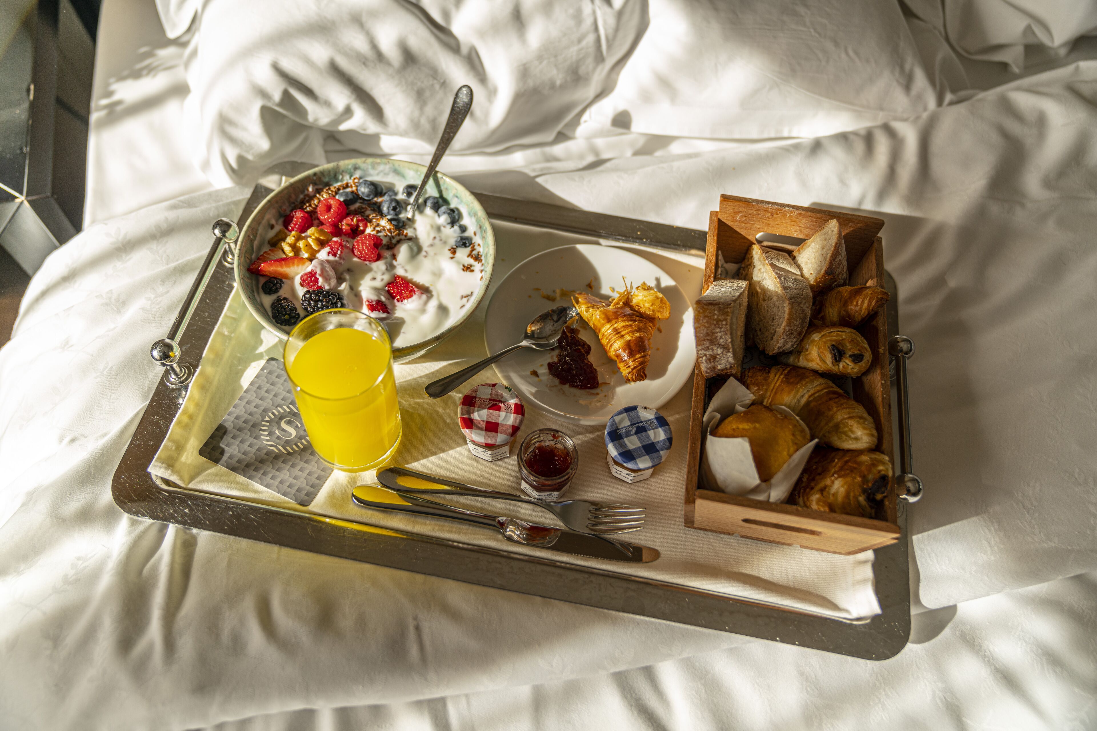 Un plateau de petit-déjeuner sur un lit, avec un bol de céréales, un verre de jus de fruits, une assiette de croissants et une boîte de pain.