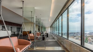 Un interior moderno con vistas panorámicas, que incluye una zona de estar con sofá y sillón colgante, un comedor con mesas y sillas, y un gran ventanal con vistas a la ciudad.