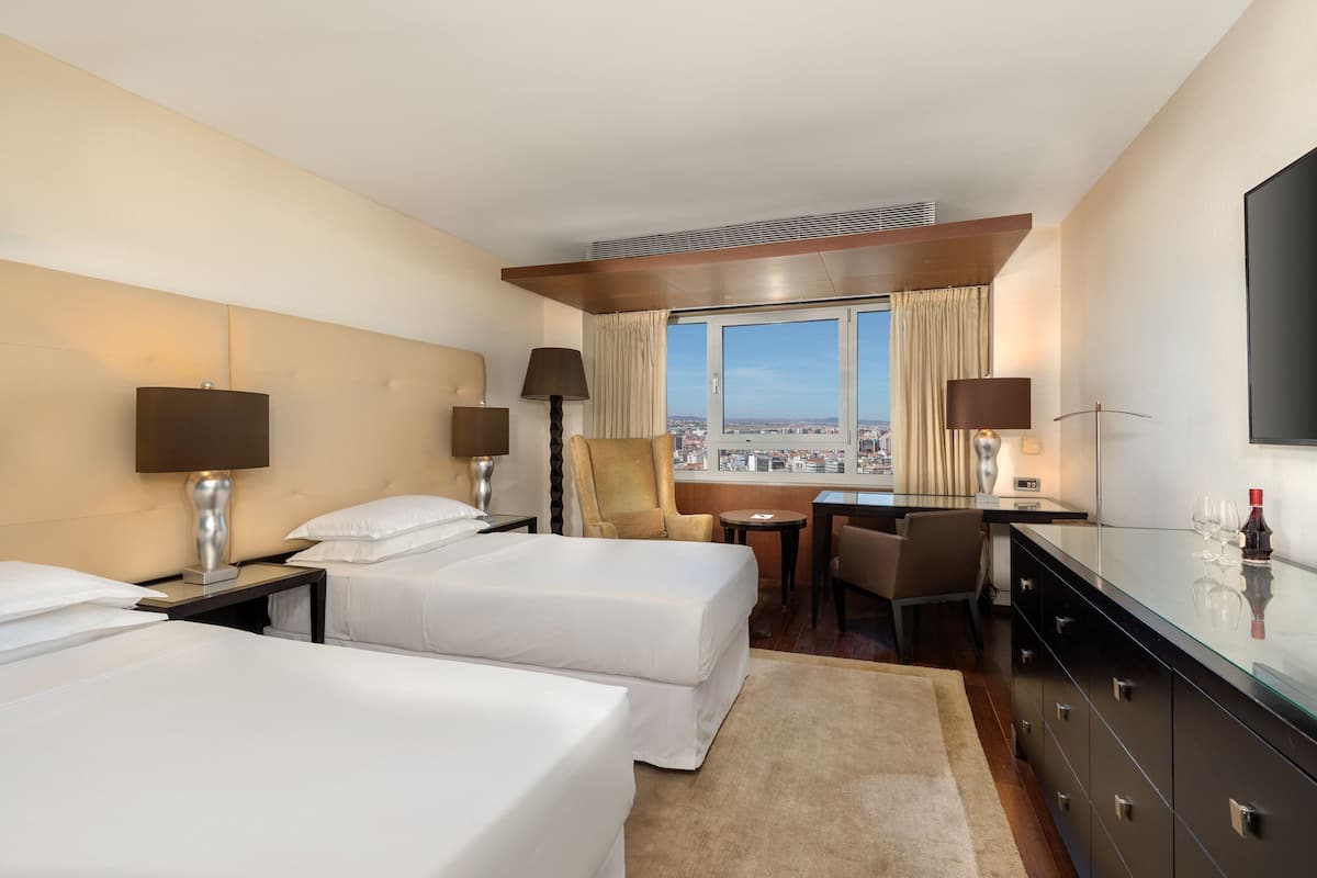 Habitación de hotel con dos camas, un escritorio, una silla, televisor y vista a la ciudad.