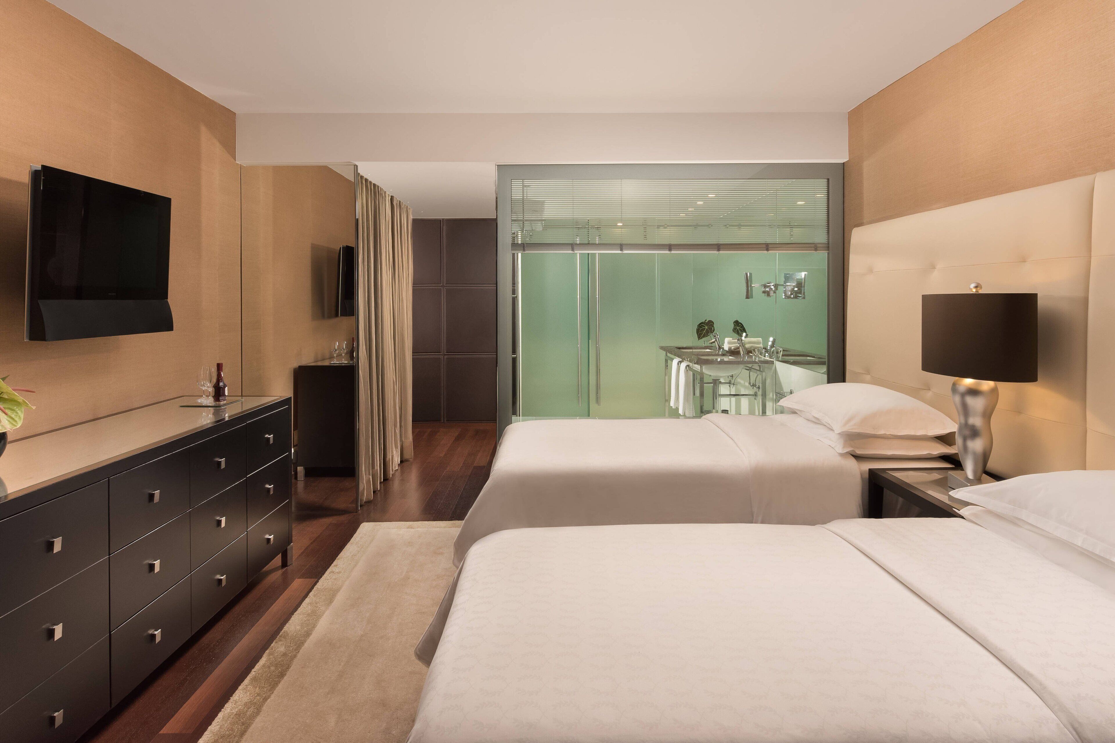 A modern hotel room with two beds, a flat-screen TV, a minibar, and a glass-enclosed bathroom.