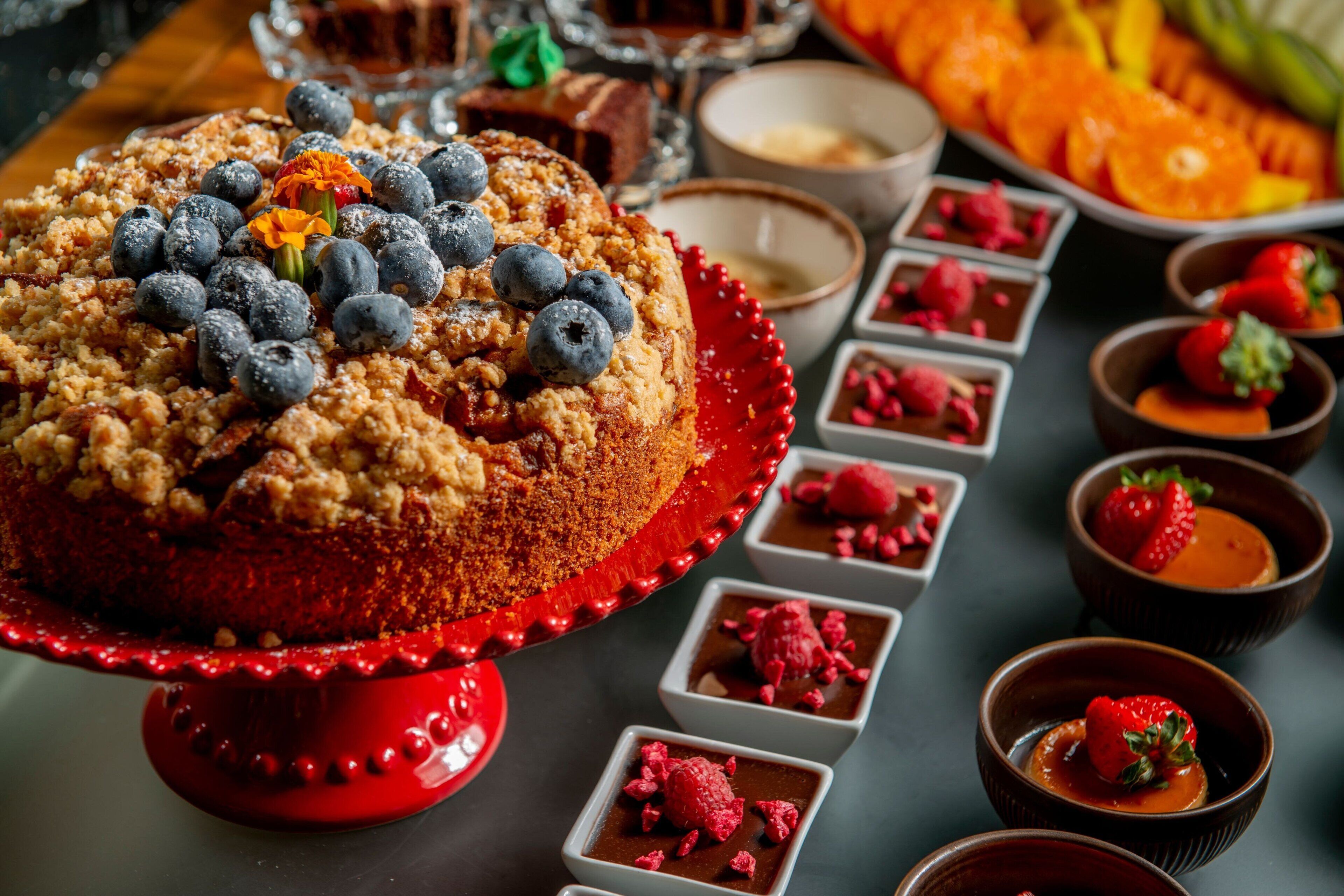 Et dessertbord med en rød kage pyntet med blåbær og en blomst, omgivet af små skåle med chokolademousse og friske bær.