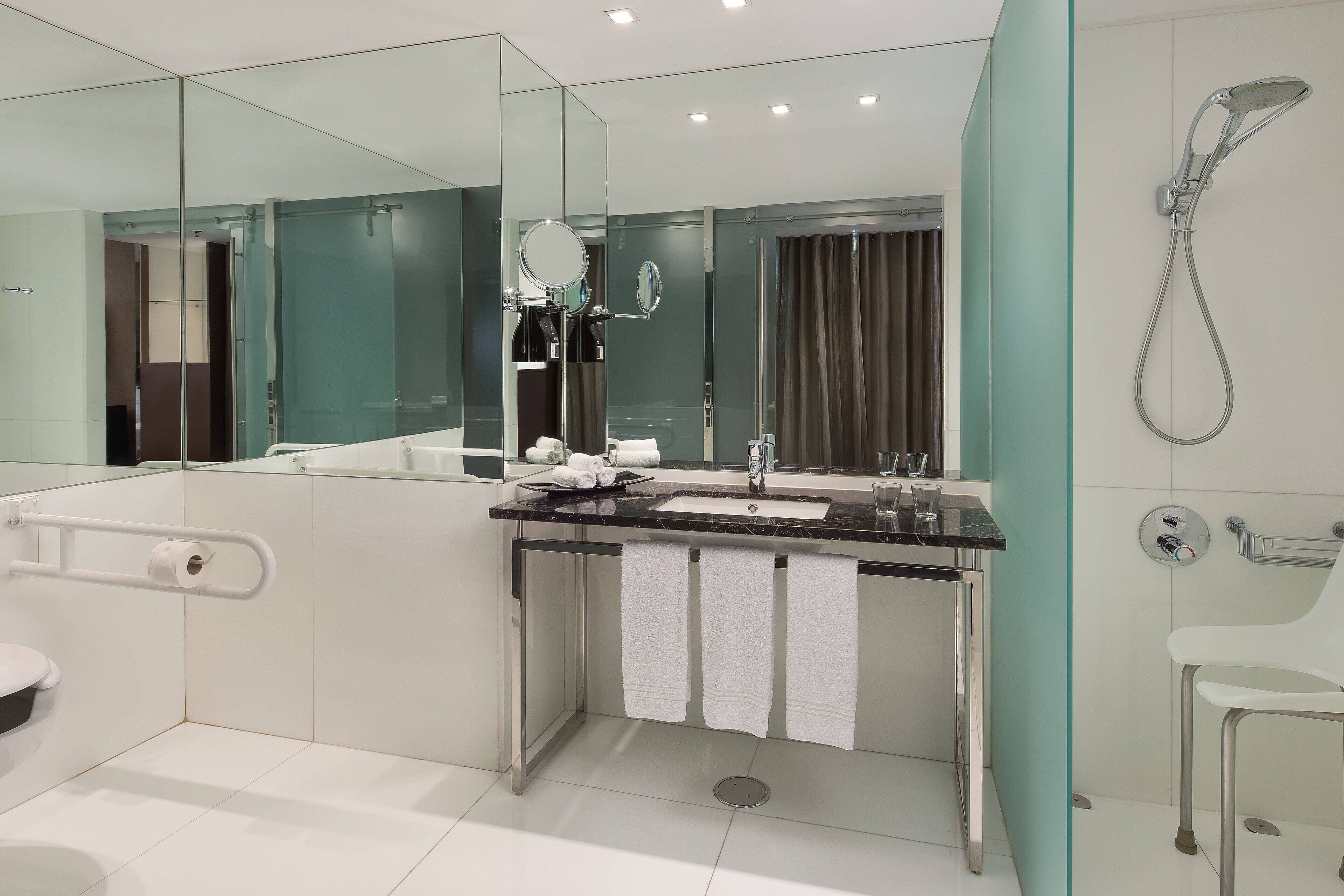 A modern bathroom with a glass shower enclosure, a large mirror, a sink with two faucets, and a toilet.