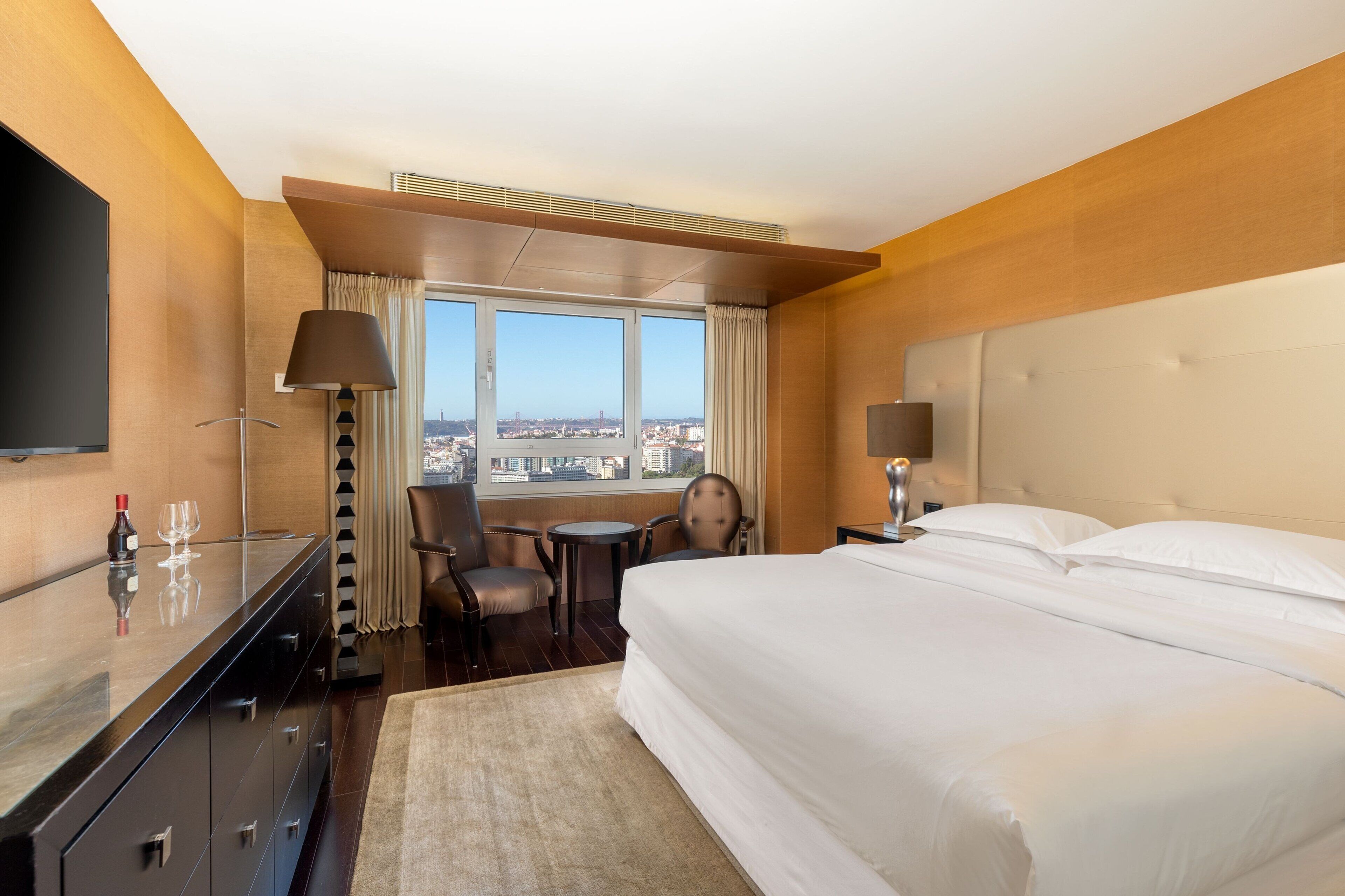 A hotel room with a large bed, a desk with a flat-screen TV, a chair, a small table, and a view of the city through the window.