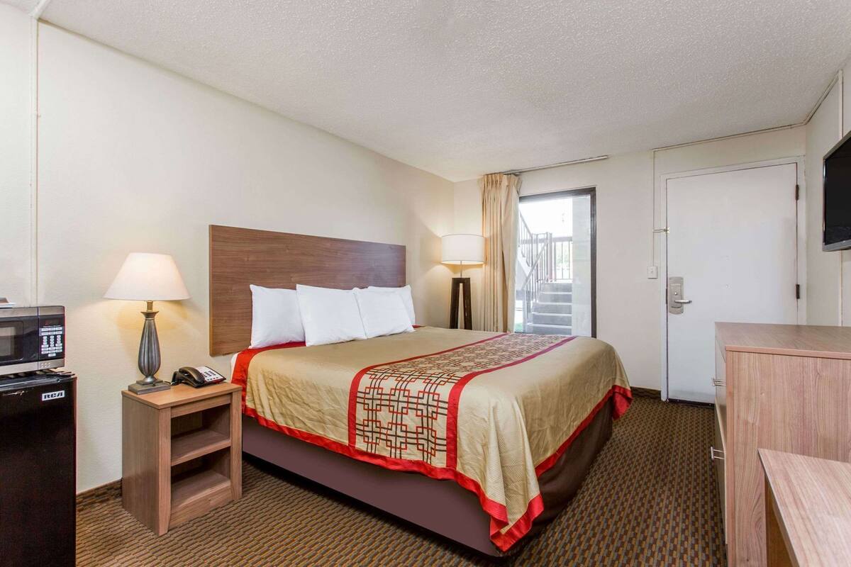 A hotel room with a bed, bedside table with a lamp, a TV, a desk with a telephone, and a microwave.