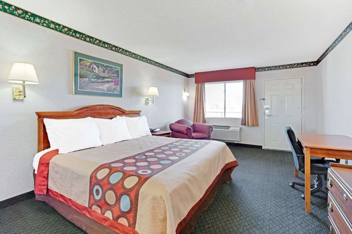 A hotel room with a bed, desk, chair, lamp, and air conditioning unit.
