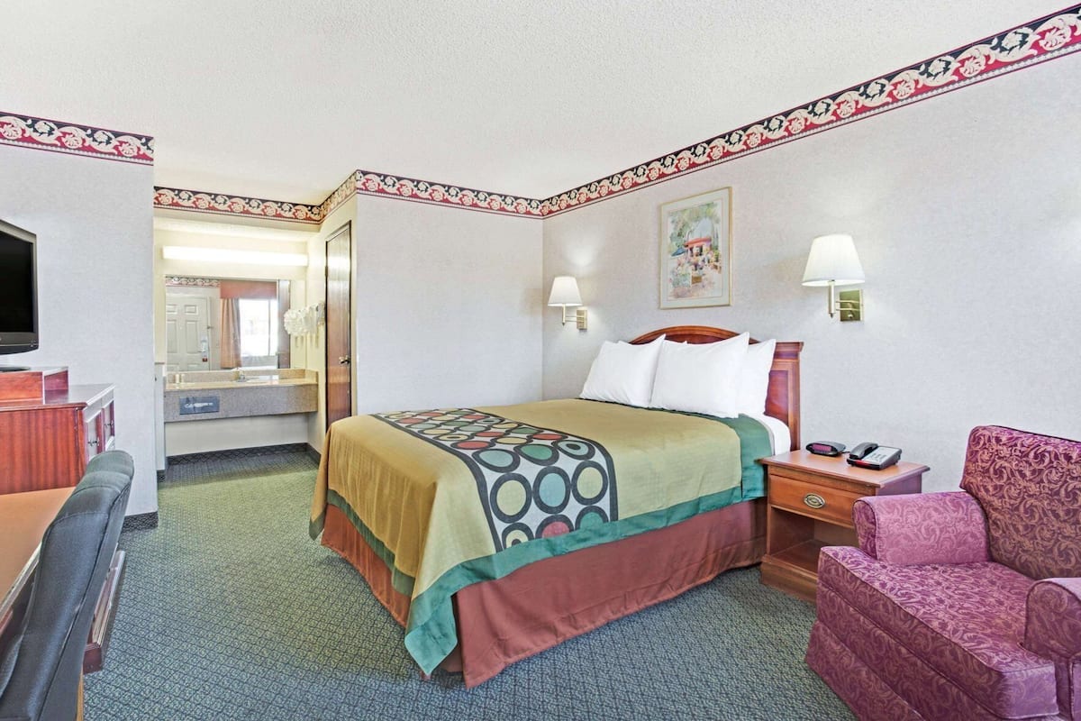 A hotel room with a bed, bedside table, lamp, chair, desk, and a television.