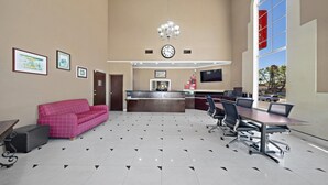 Un hall d’hôtel avec un comptoir d’accueil, un canapé rose, une table de conférence avec des chaises, une horloge et des tableaux encadrés accrochés au mur.