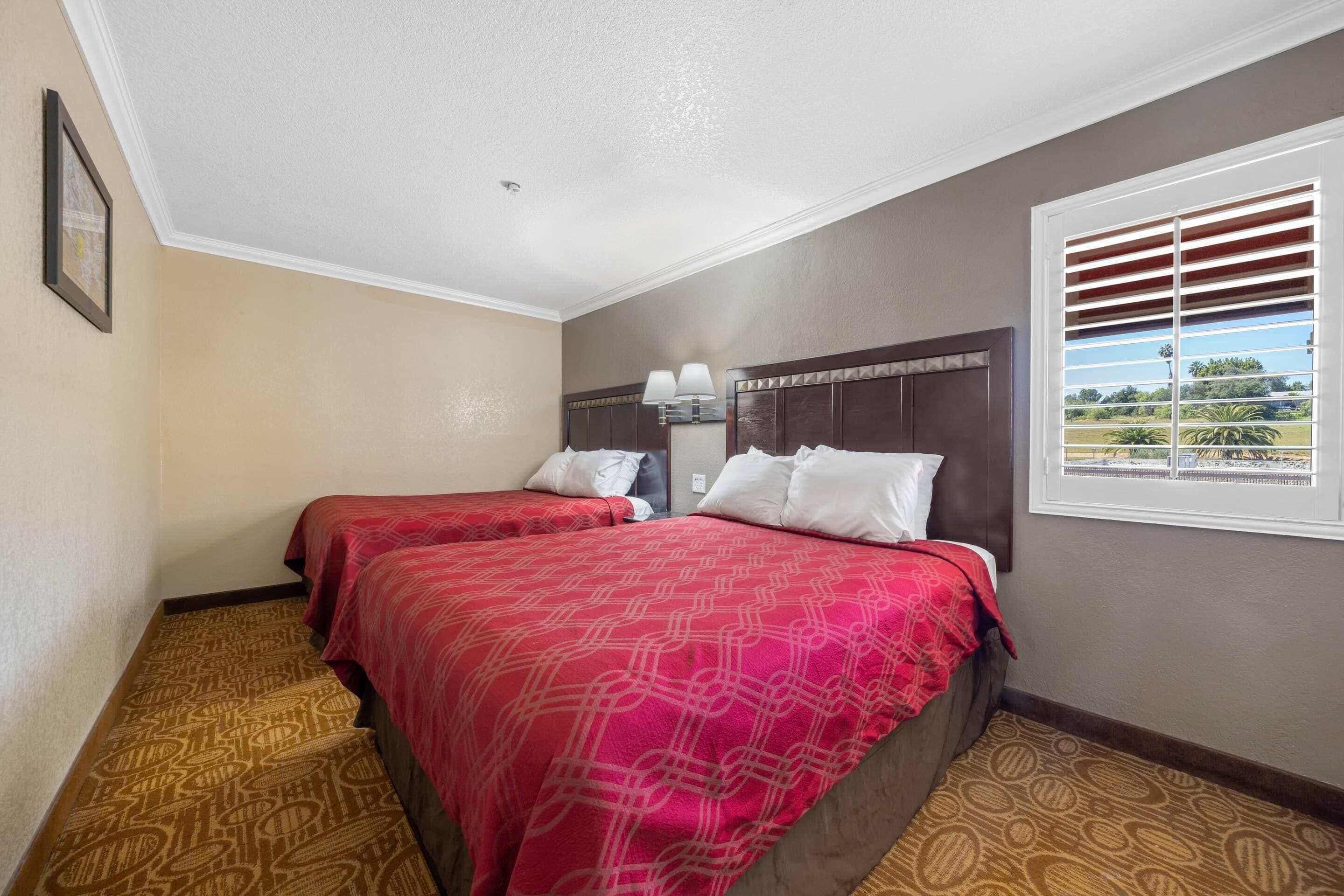 A hotel room with two beds, a window with blinds, a headboard with lamps, and a patterned carpet.