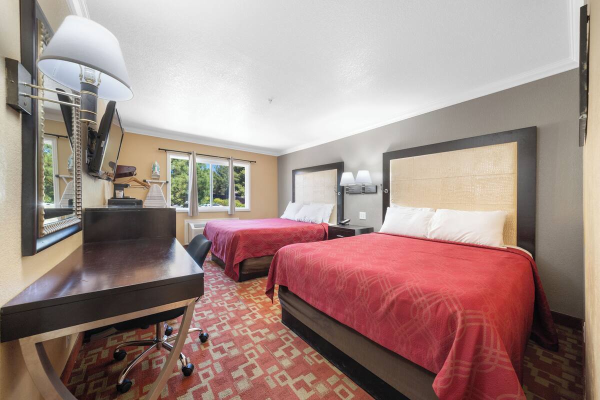 A hotel room with two beds, a desk, a lamp, and a large window.