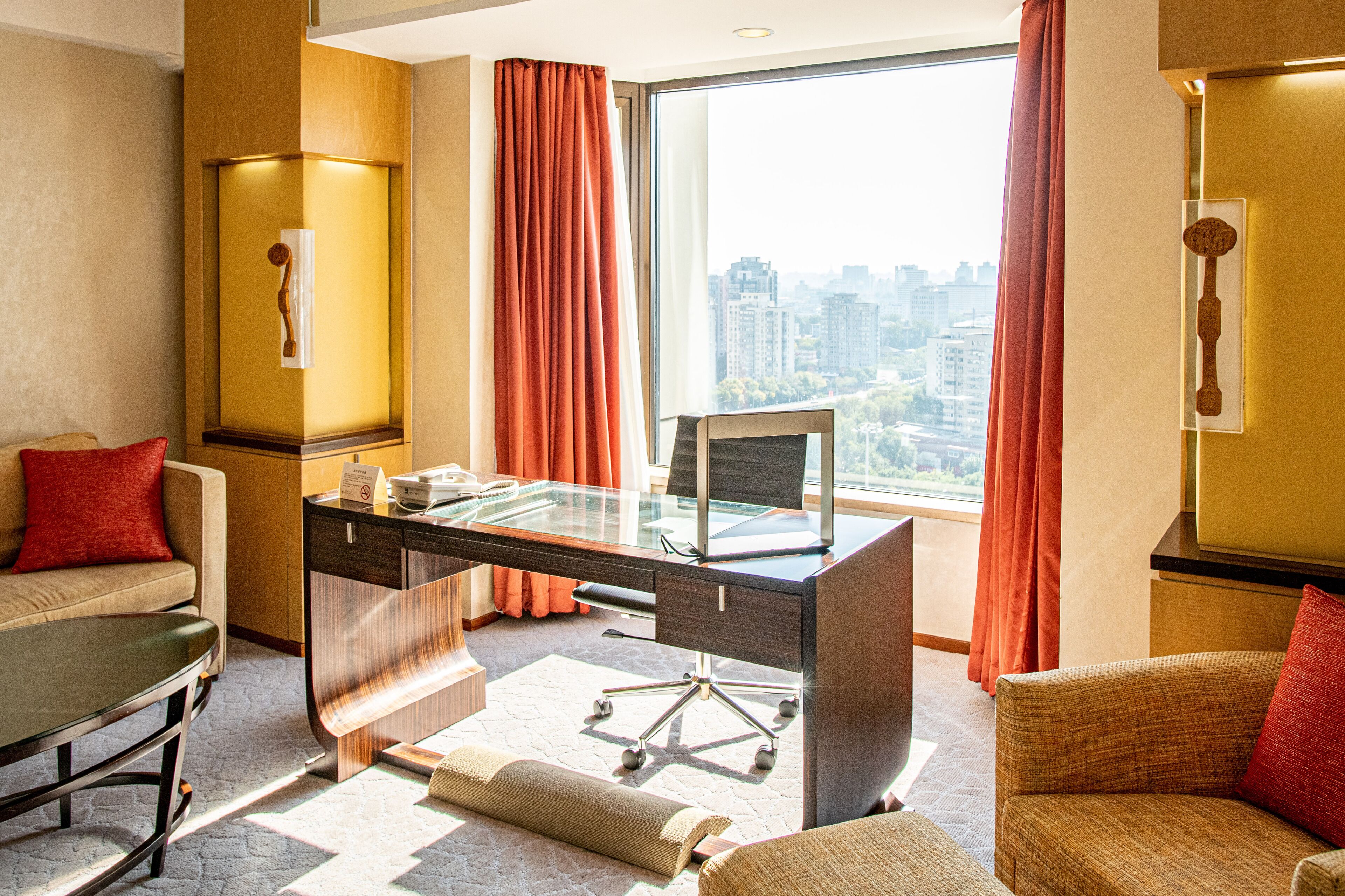 Habitación de hotel con escritorio de vidrio, sofá, silla y ventana grande con cortinas.