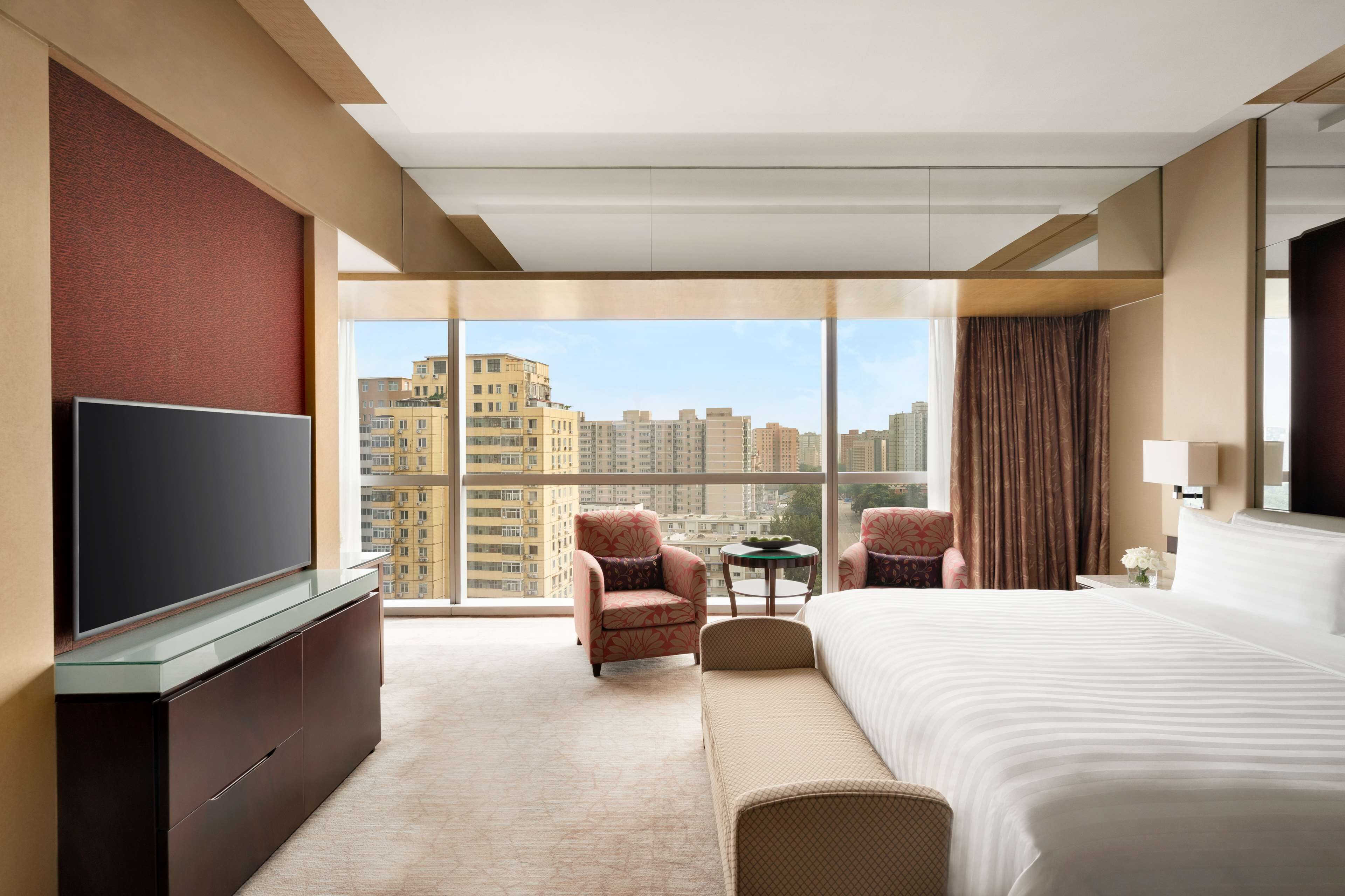 A hotel room with a large bed, a television, a seating area, and a view of the cityscape.
