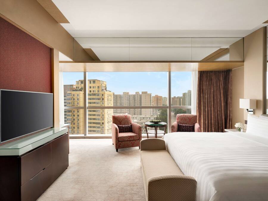 A hotel room with a large bed, a television, a seating area, and a view of the cityscape.