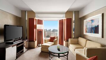 A modern hotel room with a flat-screen TV, beige sofas, a glass coffee table, and a large window offering a city view.