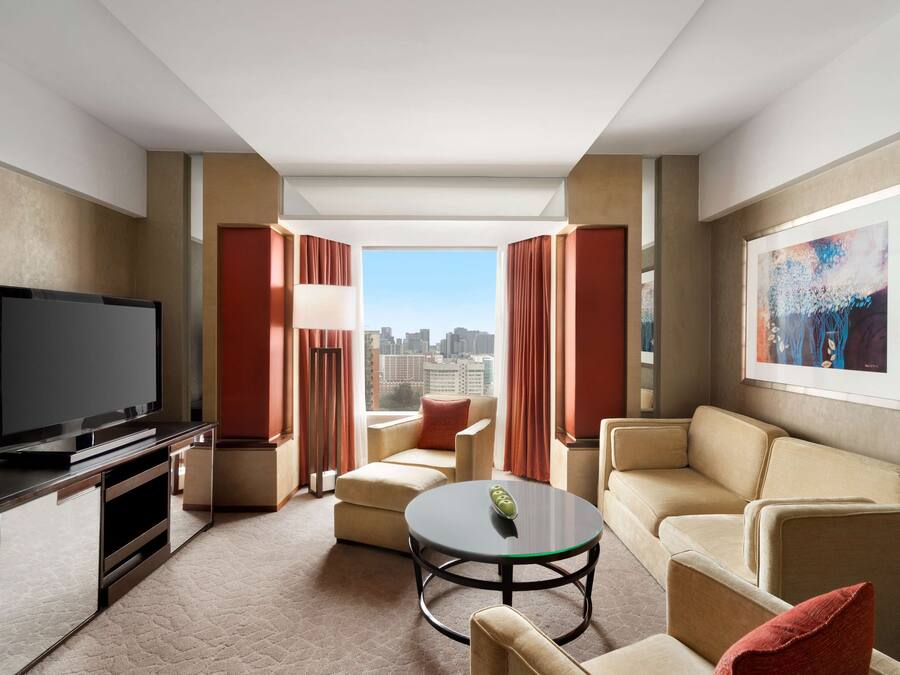 Ein modernes Hotelzimmer mit Flachbildfernseher, beigen Sofas, einem Glastisch und einem großen Fenster mit Blick auf die Stadt.