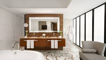 A modern bathroom with a large mirror, dual sinks, a bathtub, and a seating area.