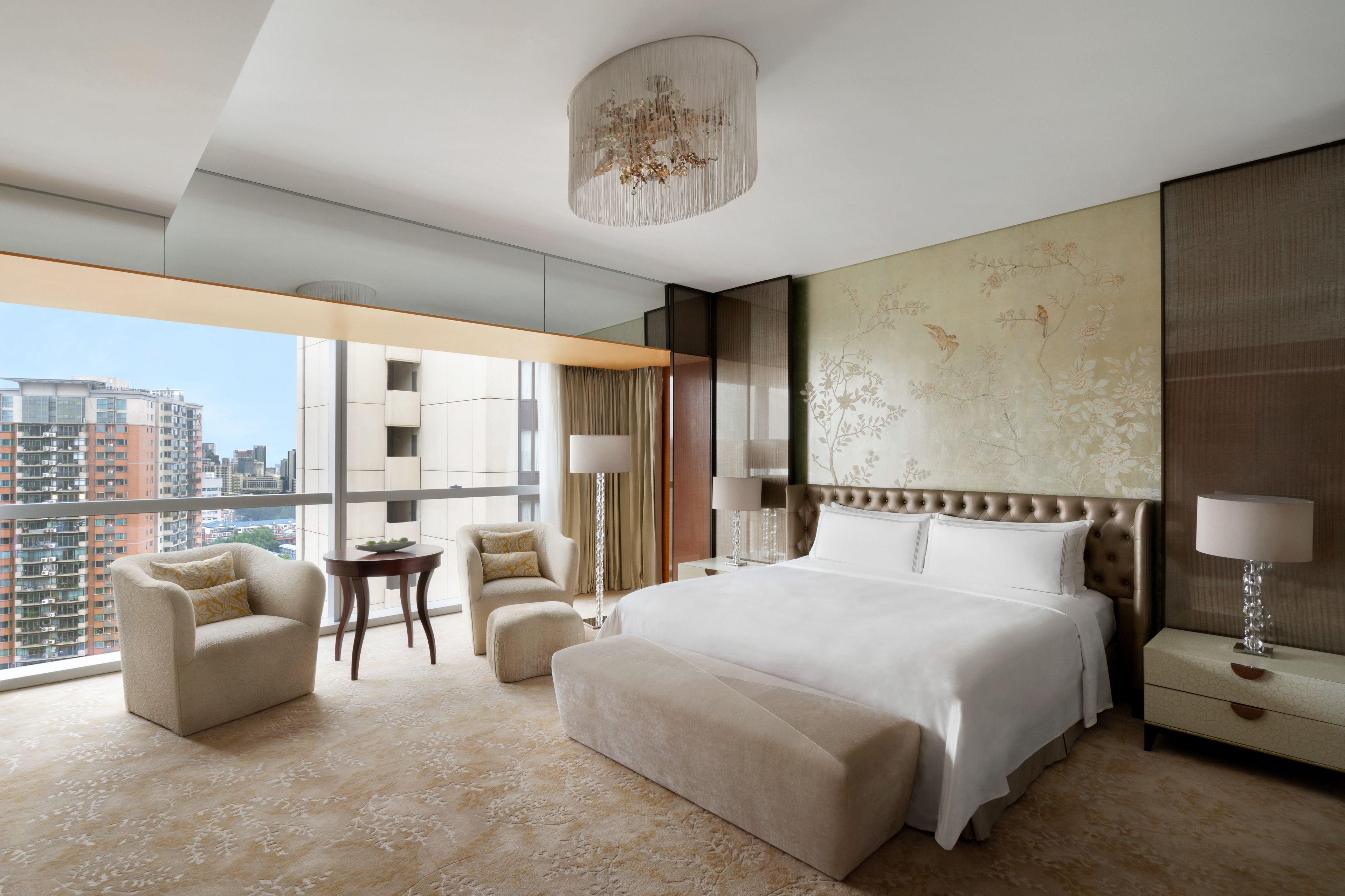 Une chambre d’hôtel moderne dotée d’un grand lit, d’un coin salon avec un fauteuil et un pouf, d’une petite table et d’une grande fenêtre donnant sur la ville.
