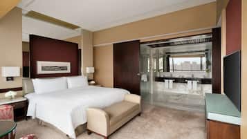 A hotel room with a large bed, a sofa, a desk with a chair, a TV, and a bathroom with a glass shower.