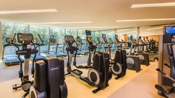 Ein Fitnessstudio mit mehreren Laufbändern und Crosstrainern, großen Fenstern mit Blick auf Bäume und einem modernen Design.