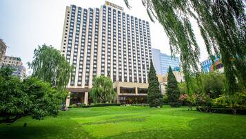 Un hotel alto con fachada moderna, rodeado de un área verde y árboles bien cuidados.