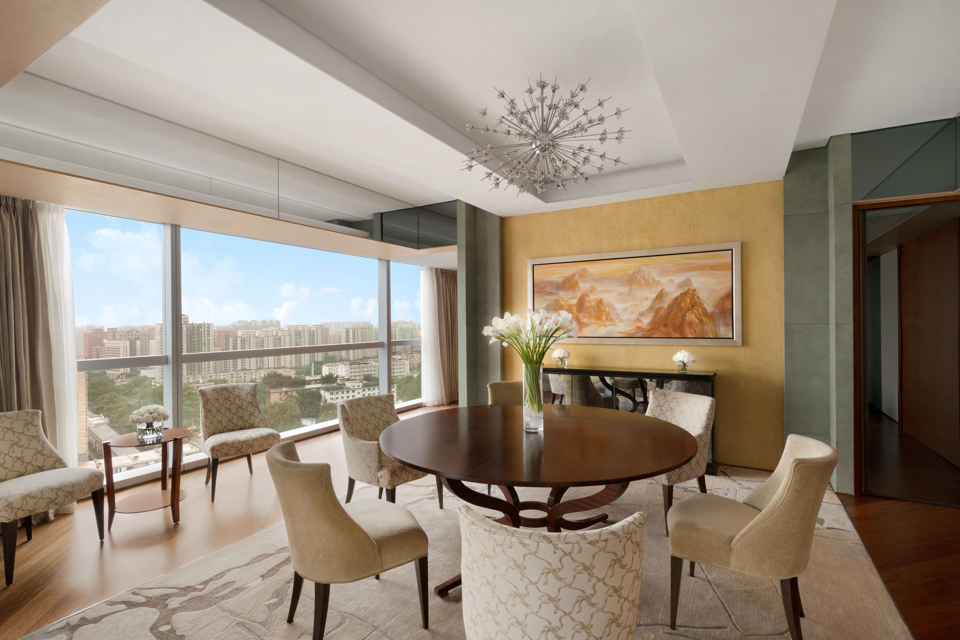 A spacious dining area with a round table and six chairs, large windows offering a city view, and a modern chandelier.