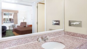 A hotel room with a bed, a desk, a chair, a mirror, a sink, and a window with curtains. - Travelodge by Wyndham Jackson (Jackson)