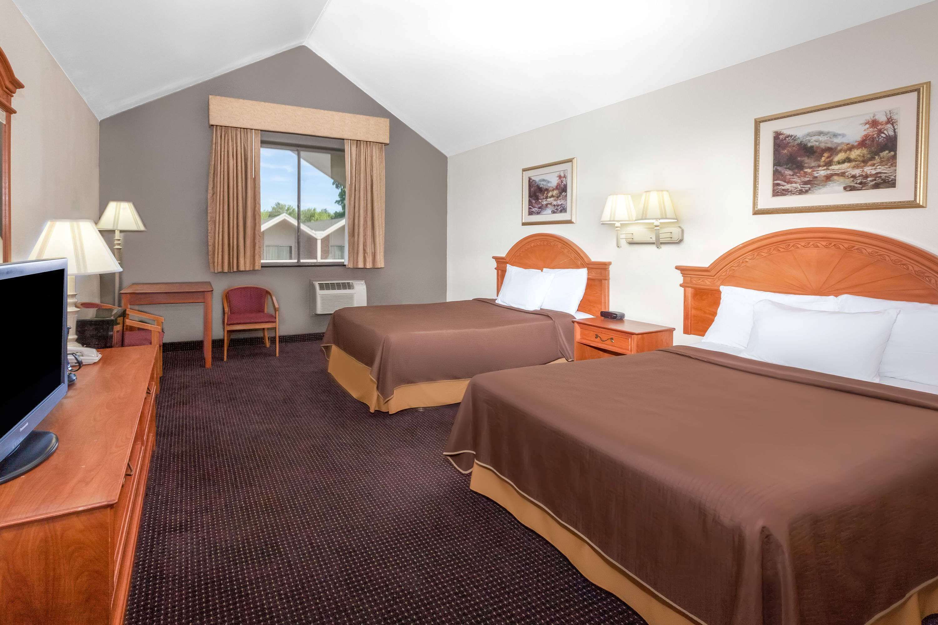 Habitación de hotel con dos camas, un escritorio con televisor, una silla, una ventana con cortinas y dos lámparas de pared.