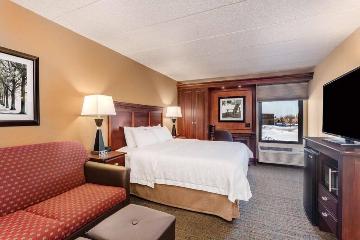 A hotel room with a bed, two bedside lamps, a television, a sofa, and a window with a view.