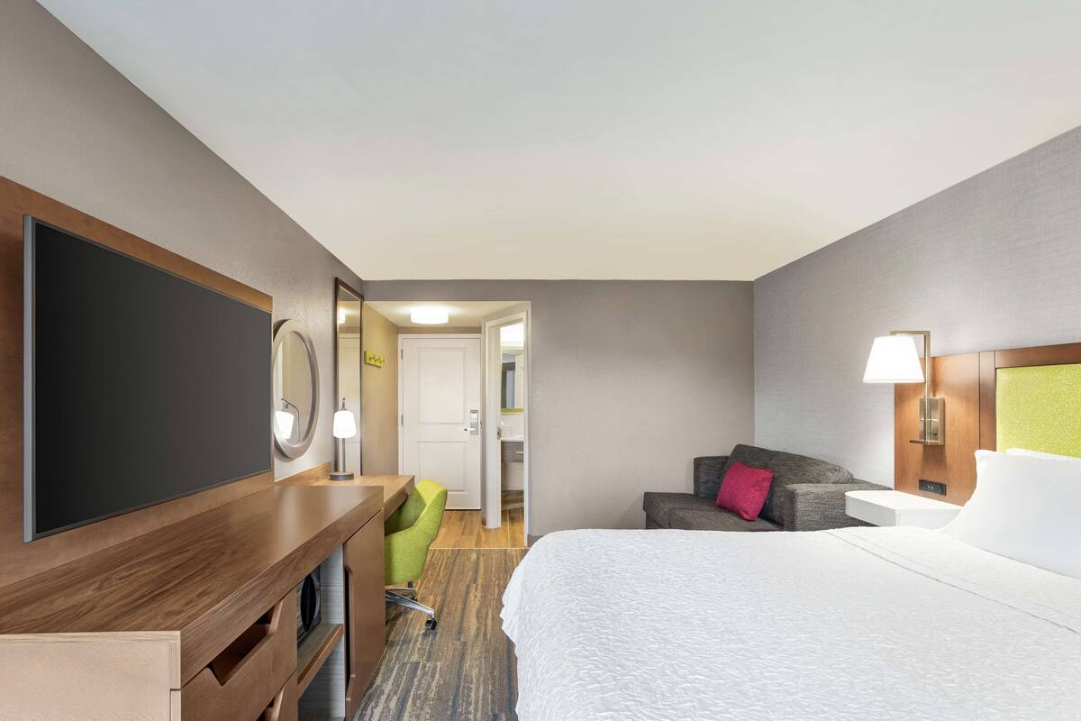 A hotel room with a bed, a desk with a flat-screen TV, a green chair, a sofa, and a door leading to a bathroom.
