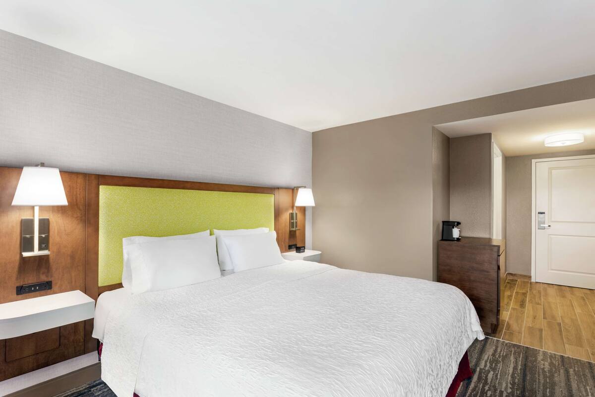 A hotel room with a large bed, two bedside tables with lamps, a wooden headboard, a small desk, and a door.