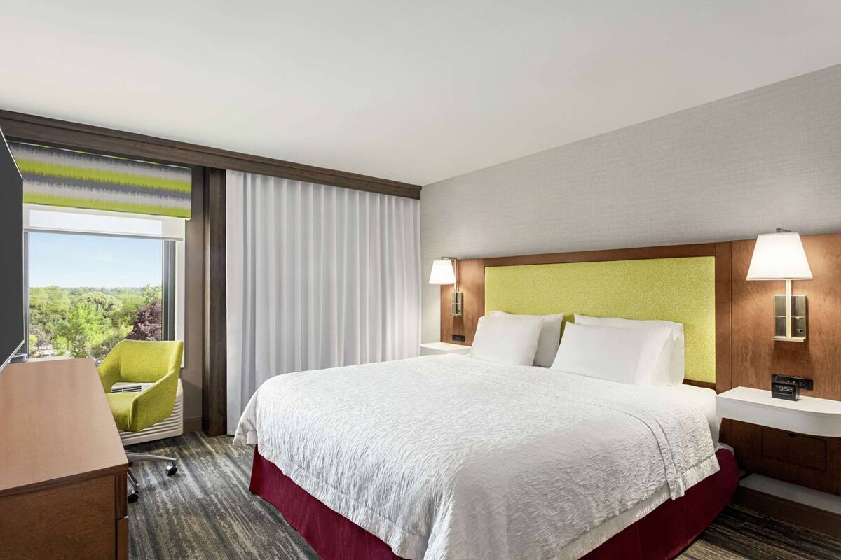A hotel room with a large bed, a desk with a chair, a window with curtains, and a view of greenery outside.