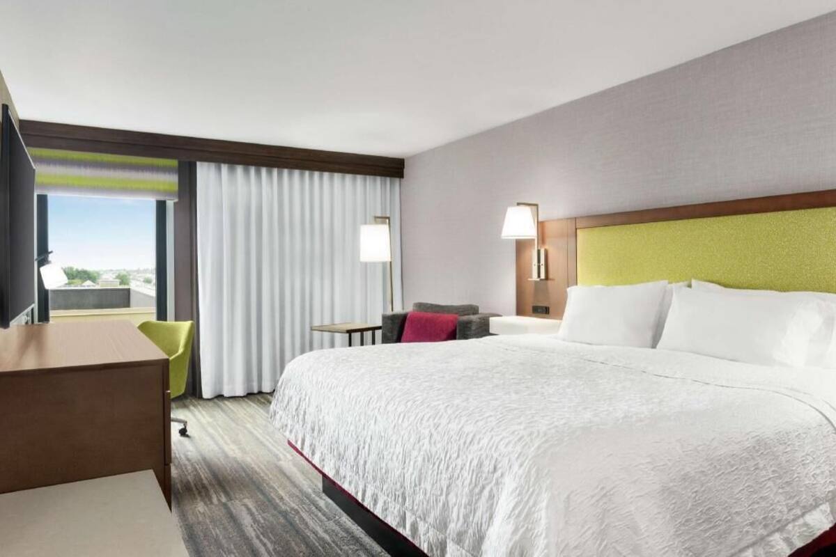 A hotel room with a large bed, a desk, a chair, a window with curtains, and a view of the city.