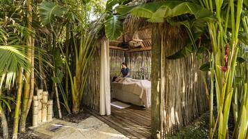 Relax in a serene garden setting with a shaded bed under a thatched roof, surrounded by lush tropical vegetation.