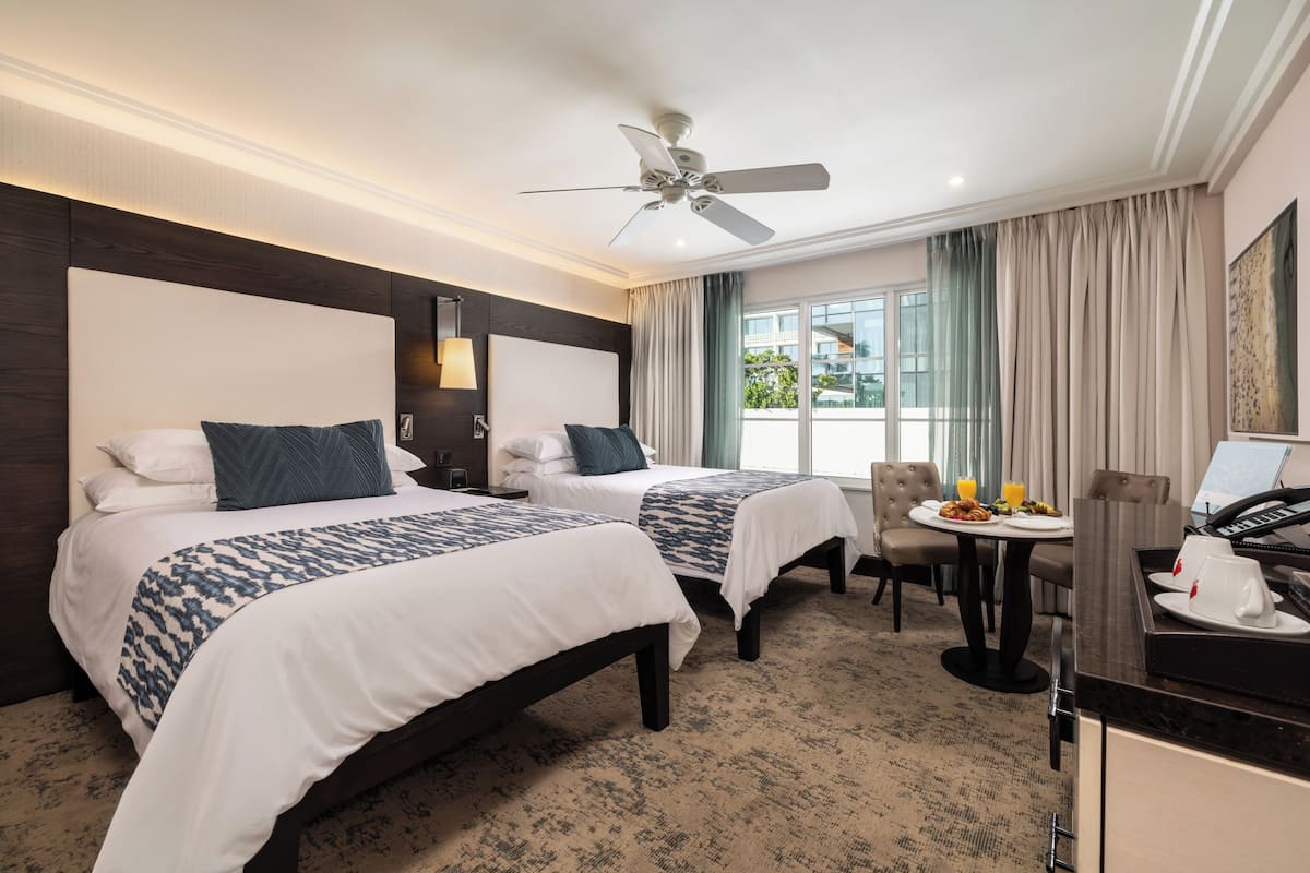 A hotel room with two beds, a dining area, a desk with a laptop, and a view of the outdoors.