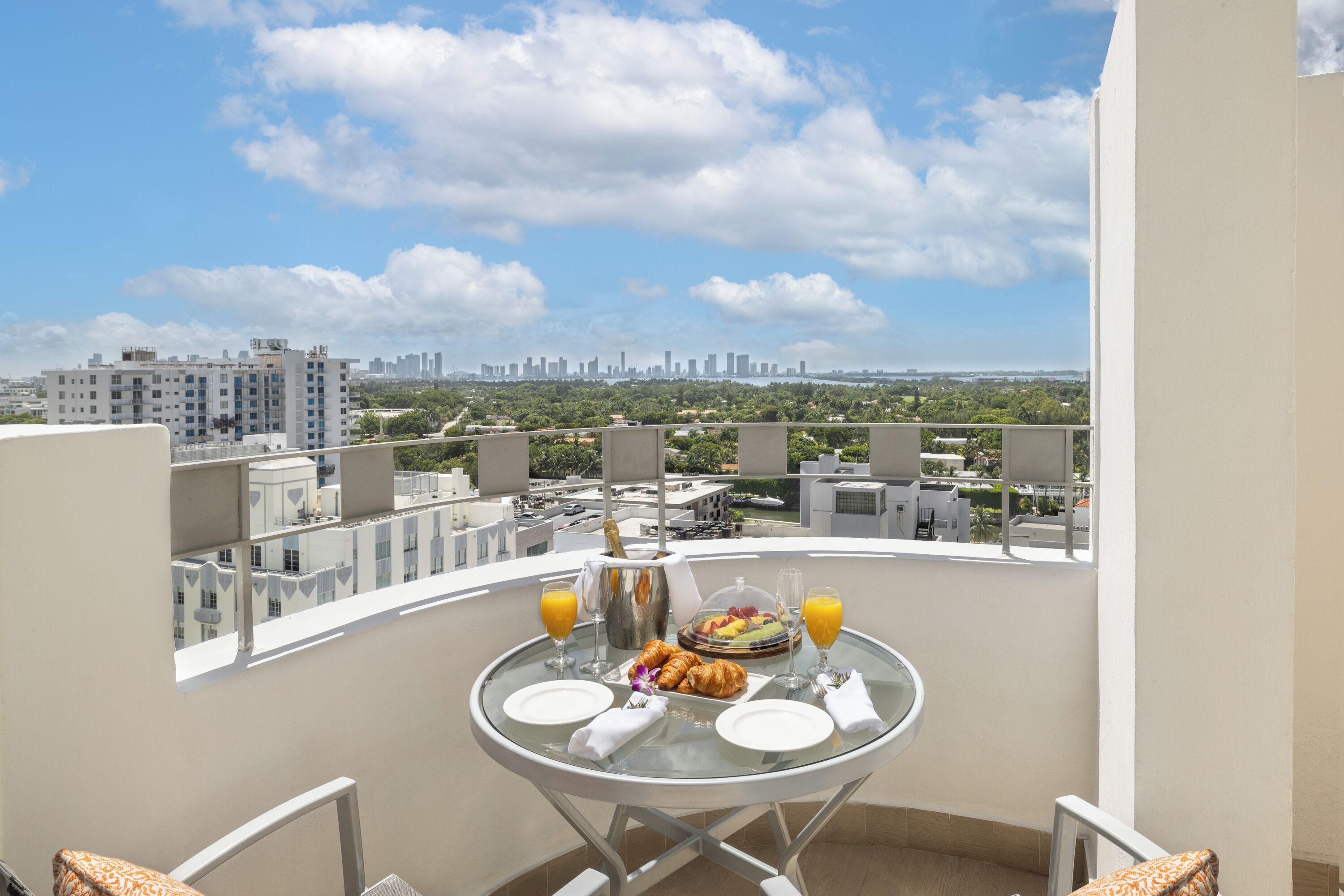 Ein Balkon mit einem Tisch, der für ein Frühstück gedeckt ist, darunter Croissants, Obst, Saft und eine Champagnerflasche. Im Hintergrund sieht man eine Stadtansicht und Grünflächen.