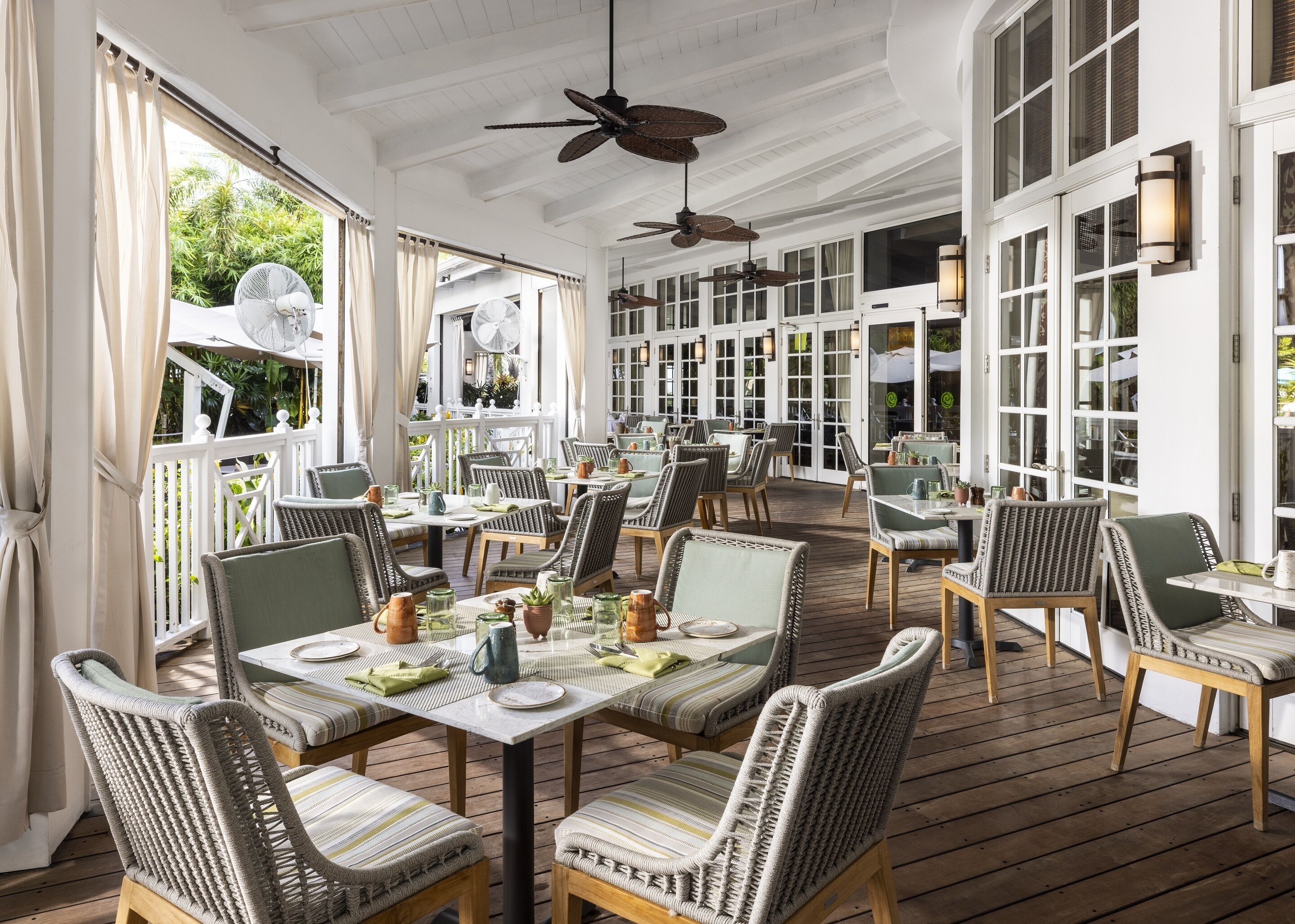 A spacious outdoor dining area with wicker chairs and tables, large windows, and ceiling fans.