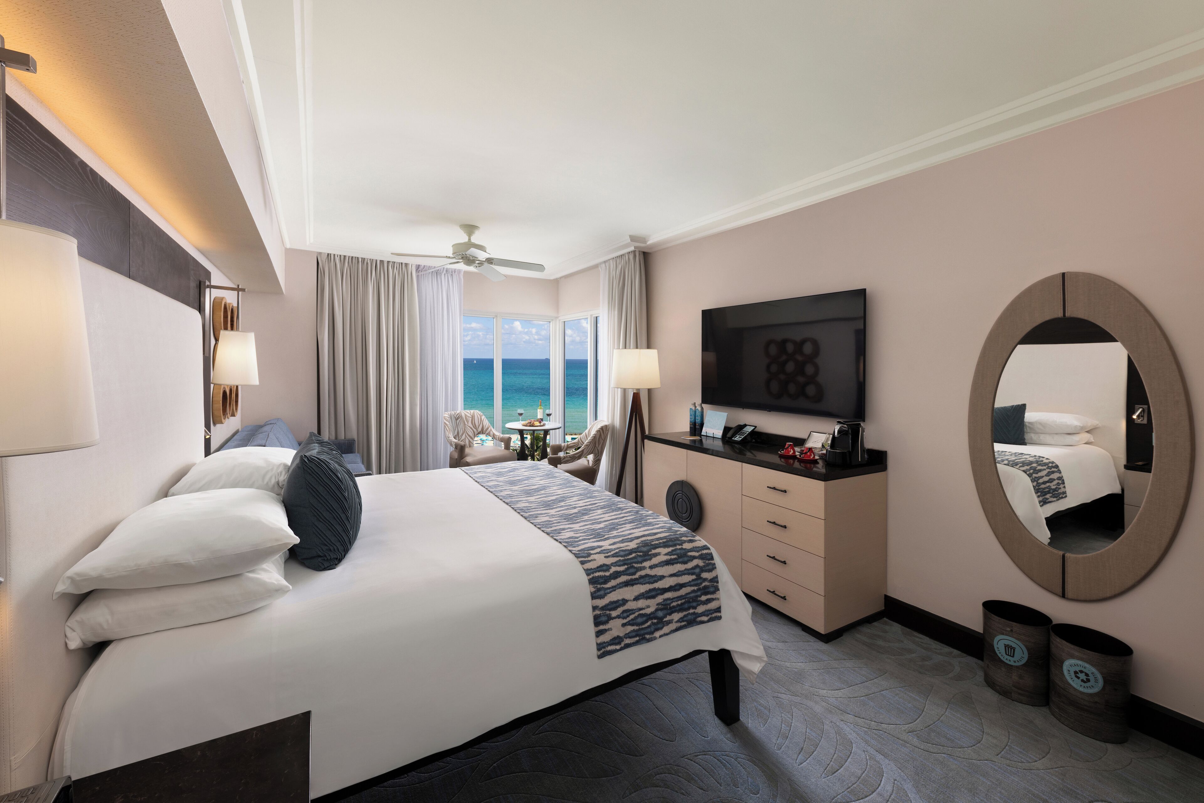 Een hotelkamer met een bed, bureau, televisie, plafondventilator en uitzicht op zee.